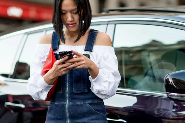 A woman in a denim dress and white off-shoulder top is looking at her phone while standing next to a car.