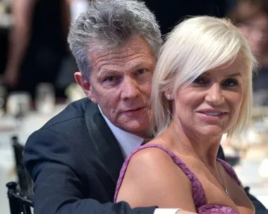 Man in dark suit and woman in lavender dress seated together at a formal event, with a blurred background of other attendees.