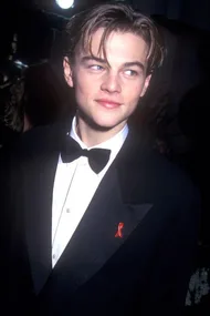 Young man in a tuxedo with a bow tie, wearing a red ribbon, looks to the side at an event.