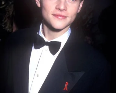 Young man in a tuxedo with a bow tie, wearing a red ribbon, looks to the side at an event.