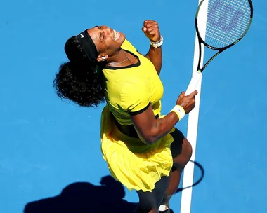 Tennis player in a yellow outfit celebrates with a clenched fist, holding a racket, on a blue court.