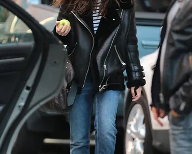 Woman in leather jacket and jeans holds an apple, walking on a cobblestone street near parked cars.