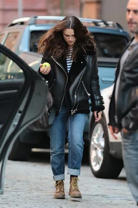 Woman in leather jacket and jeans holds an apple, walking on a cobblestone street near parked cars.