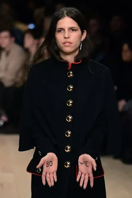 A model in a black coat at a Burberry show, holding hands up with "BO" on one palm and "WIE" on the other, paying tribute to David Bowie.