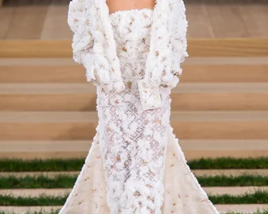 Model in a textured white gown and jacket on the Chanel Haute Couture SS16 runway with a stepped stage and grass details.