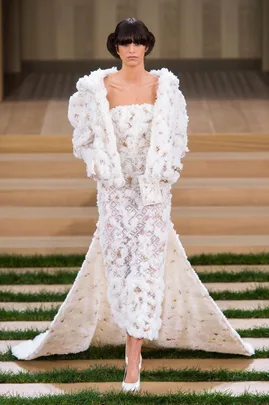 Model in a textured white gown and jacket on the Chanel Haute Couture SS16 runway with a stepped stage and grass details.