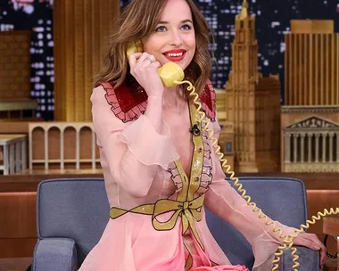 Dakota Johnson, wearing a pink and red dress, smiles while holding a yellow phone on a TV show set with a cityscape backdrop.