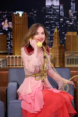 Dakota Johnson, wearing a pink and red dress, smiles while holding a yellow phone on a TV show set with a cityscape backdrop.