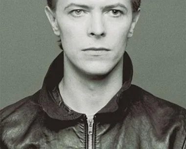 Monochrome photo of David Bowie wearing a leather jacket, looking directly at the camera with a neutral expression.