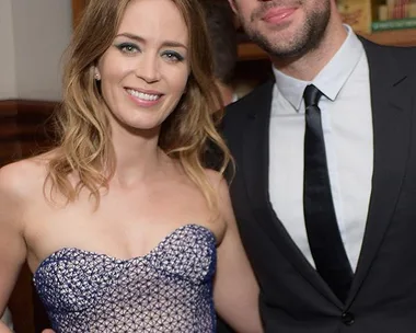 Emily Blunt and John Krasinski pose together, smiling at an event, with Blunt wearing a strapless dress and Krasinski in a suit.