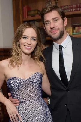 Emily Blunt and John Krasinski pose together, smiling at an event, with Blunt wearing a strapless dress and Krasinski in a suit.