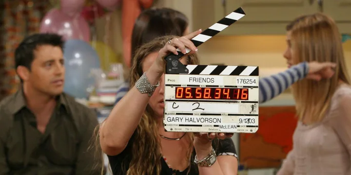 Behind-the-scenes of Friends with a clapperboard; Joey and Rachel are visible in the background.