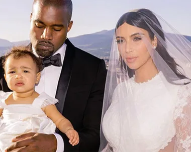 A man in a tuxedo, a woman in a wedding dress with a veil, and a baby in a white dress, posing outdoors with a scenic background.