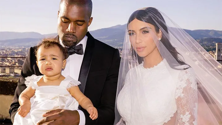 A man in a tuxedo, a woman in a wedding dress with a veil, and a baby in a white dress, posing outdoors with a scenic background.