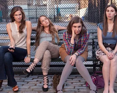 Four women sit on a bench in a park, each with distinct styles and expressions, from the TV show "Girls."