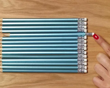 A hand with red nail polish adjusts a misaligned pencil among a row of perfectly aligned pencils on a wooden surface.