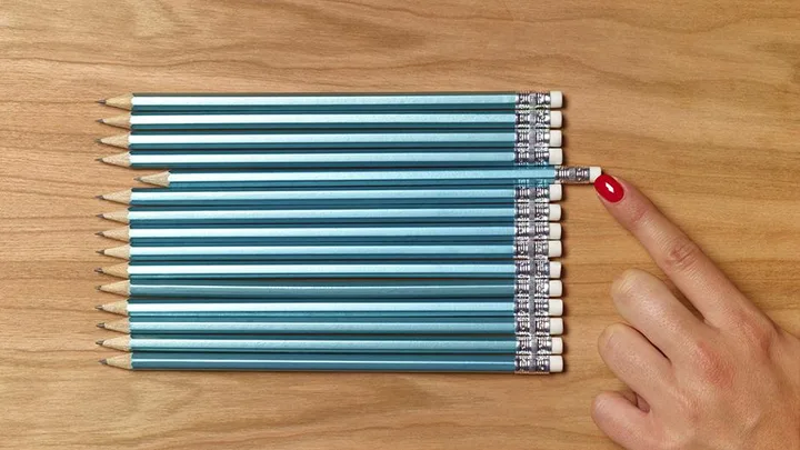 A hand with red nail polish adjusts a misaligned pencil among a row of perfectly aligned pencils on a wooden surface.