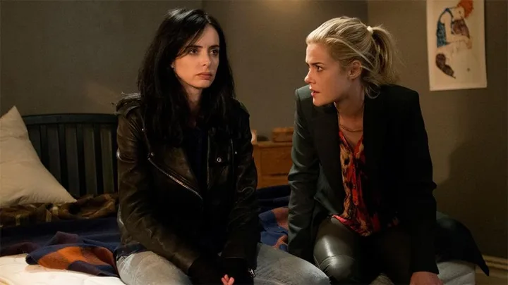 Two women, one with dark hair in a leather jacket and the other blonde, sit on a bed in a dimly lit room.