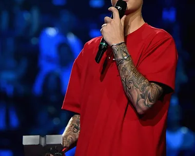 Justin Bieber, in a red shirt and gray cap, holds a mic and an award on stage, against a blue-lit background.