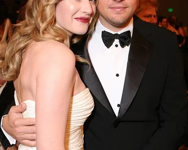 Kate Winslet and Leonardo DiCaprio pose together at an event; Kate in a white dress, Leonardo in a black tuxedo.