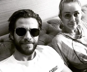 Black and white photo of two people sitting on a couch, the man wearing sunglasses and the woman smiling broadly.