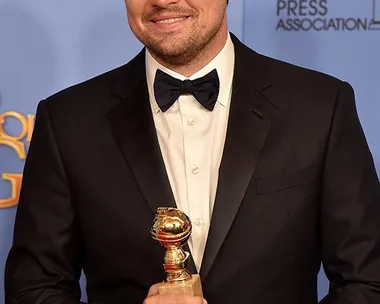 Leonardo DiCaprio in a black suit holding a Golden Globe award, with a smile on his face in front of a blue background.