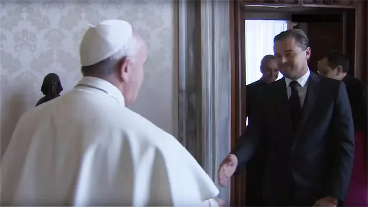 A man in a suit greets a person dressed in white robes, in what appears to be a formal setting with other people in the background.