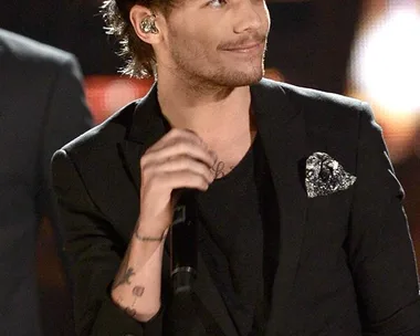 Louis Tomlinson wearing a black suit and holding a microphone on stage, with a slight smile.