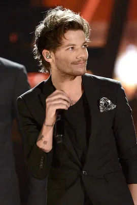 Louis Tomlinson wearing a black suit and holding a microphone on stage, with a slight smile.