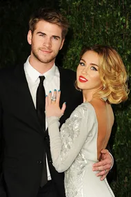 "Miley Cyrus in a white dress showing her engagement ring, posing with Liam Hemsworth in a black suit at an event."