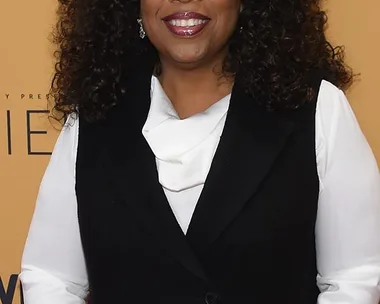 A woman with curly hair smiling, dressed in a black vest over a white blouse, standing in front of an orange background with text.