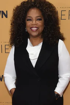 A woman with curly hair smiling, dressed in a black vest over a white blouse, standing in front of an orange background with text.