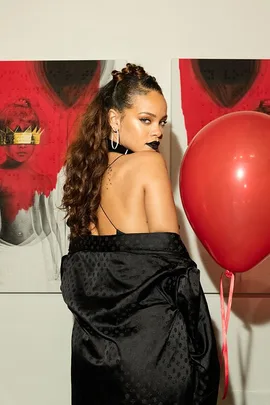 Rihanna in a black outfit holding a red balloon, standing in front of artwork from her album "Anti" with a red and black theme.