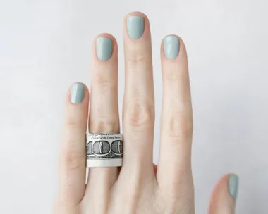 A hand with light blue nail polish wearing a rolled-up $100 bill as a ring on the ring finger.