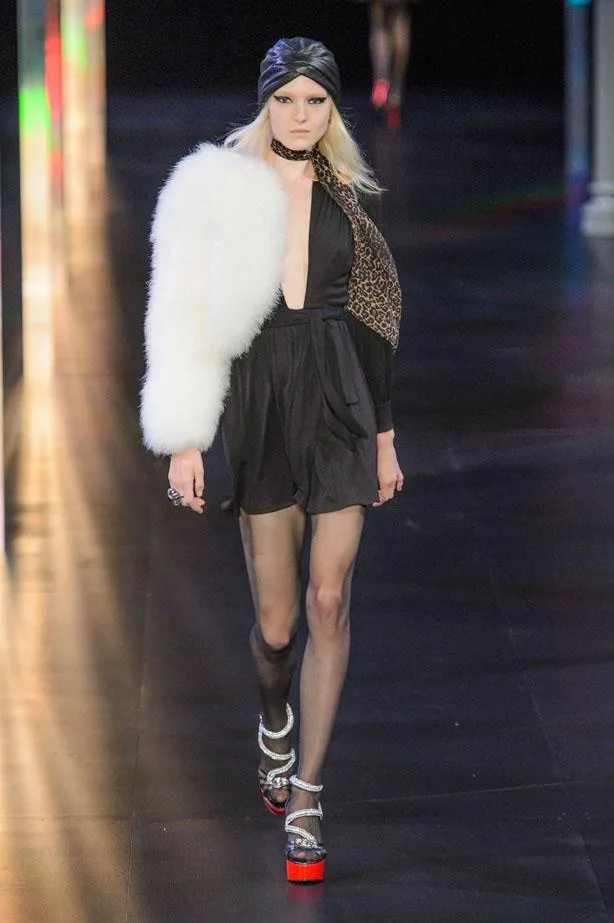 Model on runway wearing black dress, white fur coat, leopard print scarf, black turban, and ankle-strap platforms.