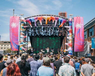 Outdoor concert with a crowd facing a colorful stage with a band performing, large decorations, and banners on either side.