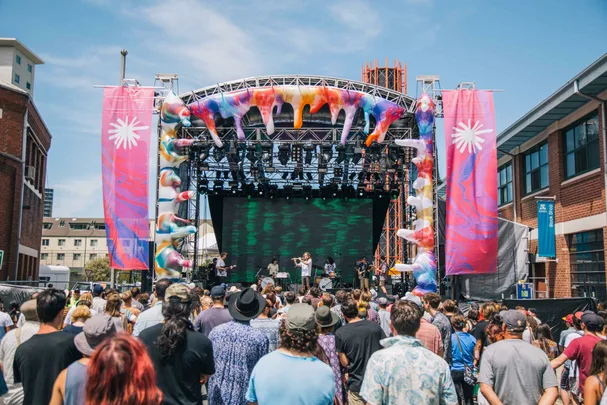 Outdoor concert with a crowd facing a colorful stage with a band performing, large decorations, and banners on either side.