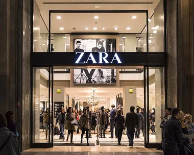 A busy Zara store entrance with shoppers inside and outside, under a lit Zara sign and black-and-white fashion ads.