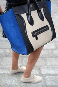 Person carrying large blue and beige handbag while wearing white sneakers on cobblestone pavement.