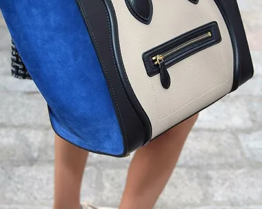 Person carrying large blue and beige handbag while wearing white sneakers on cobblestone pavement.