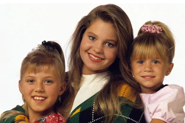 Three young girls from "Full House" smiling at the camera; middle one with long hair, others with ponytails.