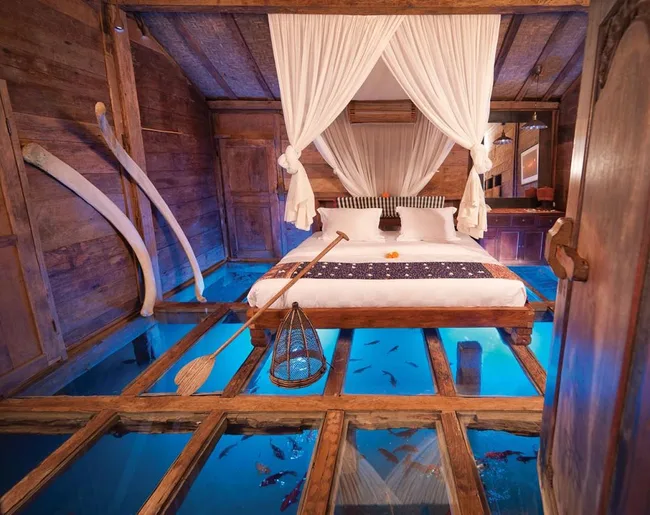 Rustic bedroom with glass floor revealing water and fish, wooden beams, and a canopied bed with white drapes.