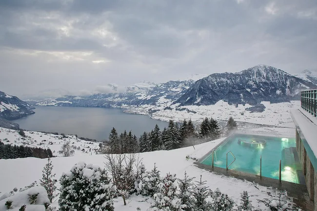 Snow-covered landscape with a steaming outdoor pool overlooking a serene lake and mountains under a cloudy sky.