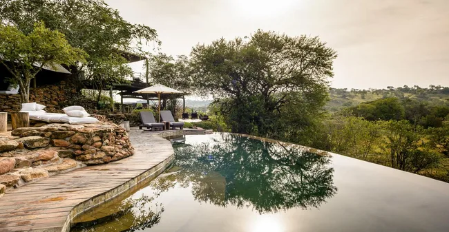 Infinity pool with lounge chairs and umbrellas, surrounded by lush greenery under a soft, cloudy sky.