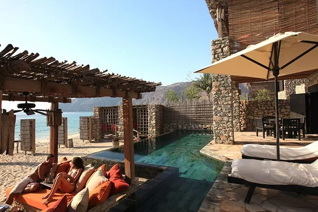 A couple relaxes on a terrace near a private pool and sun loungers, overlooking a beach and mountains.