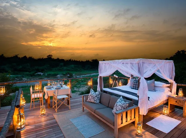 Outdoor bed with canopy and table beside it on wooden deck at sunset, surrounded by lanterns.