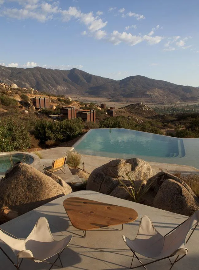Outdoor patio with two chairs and a table overlooking an infinity pool, mountainous landscape in the background.