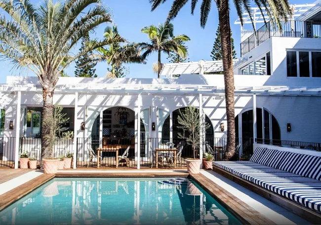 Luxurious outdoor pool area with striped loungers, palm trees, and a white modern house in the background.