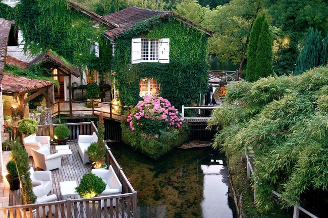 Lush garden with ivy-covered houses, wooden patio with chairs by a stream, surrounded by trees and pink flowers.