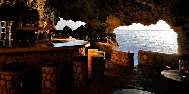 Bar inside a cave overlooking the ocean at sunset, with stone furniture and a cozy atmosphere.